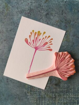 Ręcznie rzeźbiona pieczątka z motywem delikatnego kwiatu, odbita w różowo-żółtych kolorach na białym papierze.